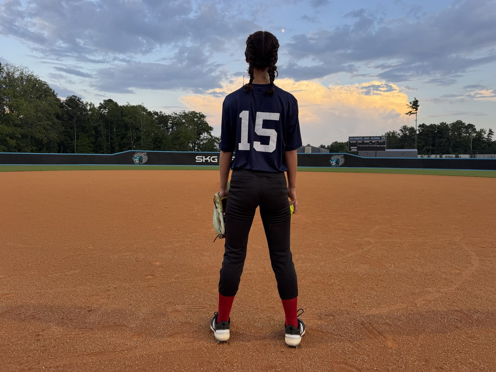 An ORIGIN athlete in a navy shirt numbered 15 stands on the infield at dusk, glove in one hand and a ball in the other.